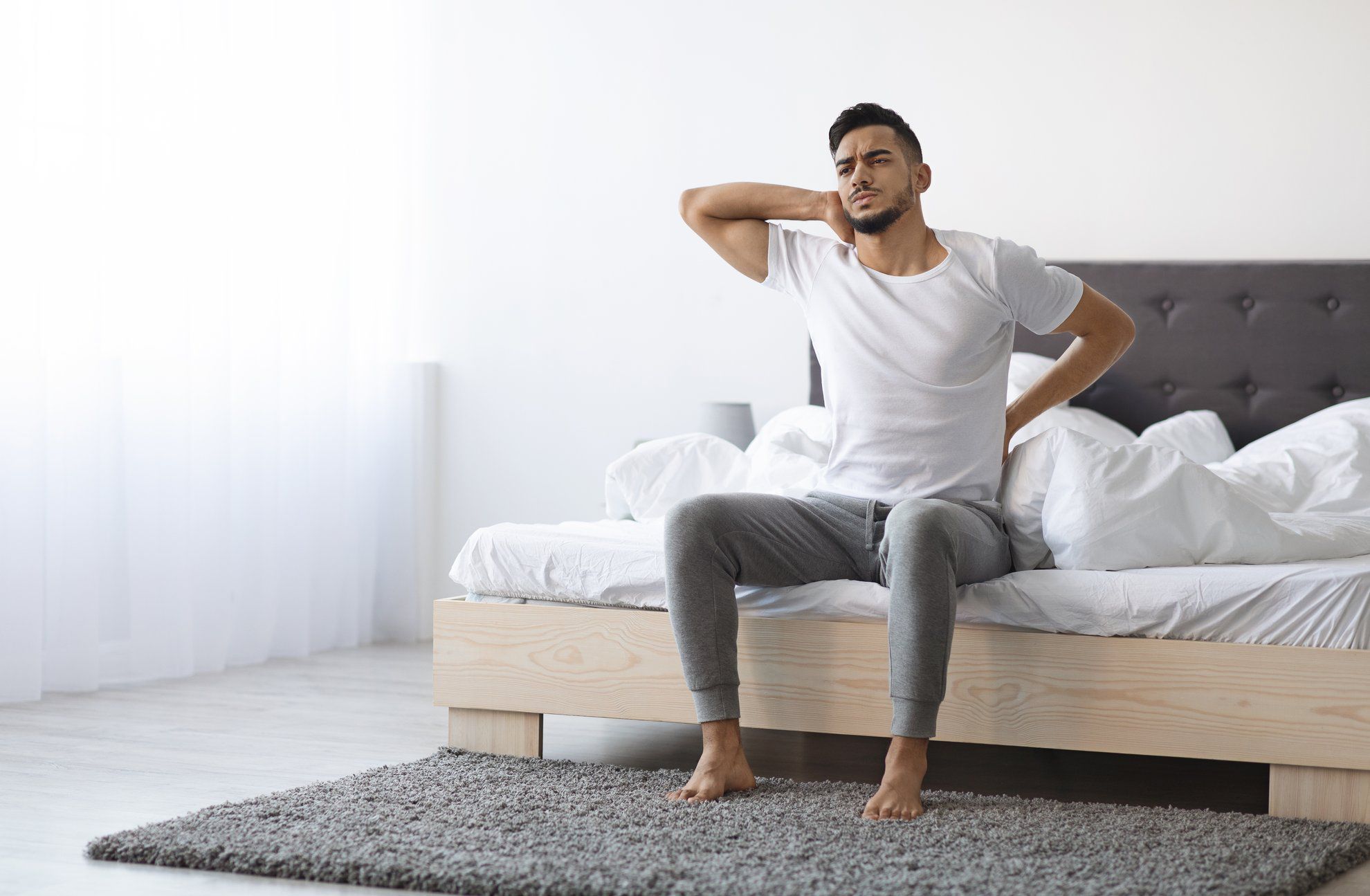 Uncomfortable Bed. Arab Man Waking Up With Neck And Back Pain, Young Middle Eastern Male Suffering Backache While Sitting In Bedroom At Home, Frowning And Massaging Aching Zones, Copy Space