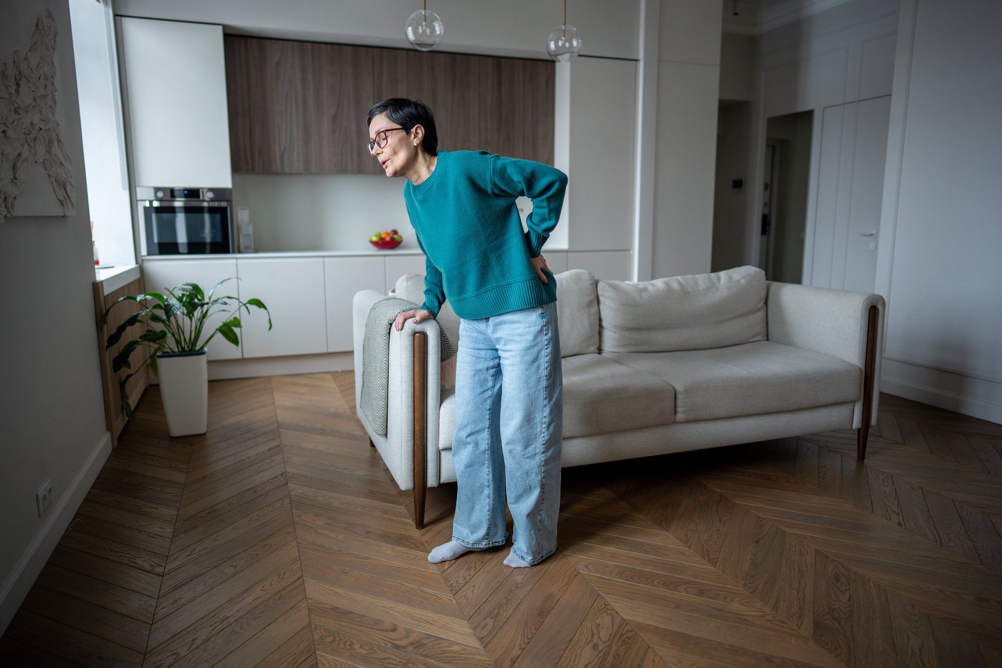 Suffering sad middle aged woman bending in pain, clutching sofa due to back injury. Lower back discomfort, spinal muscle strain, senior female health issues at home, herniated disc, posture problem