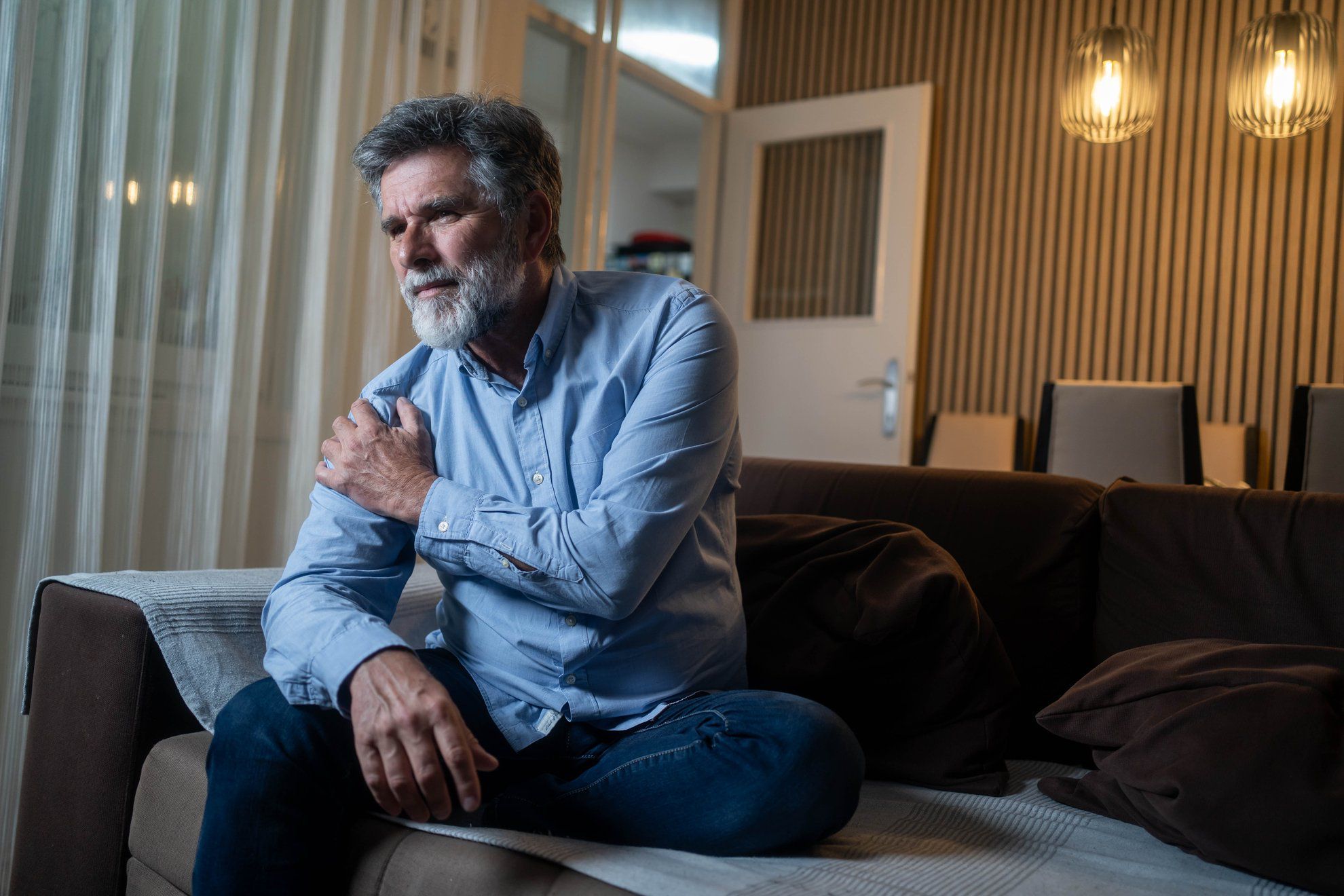 Mature man experiencing shoulder pain while sitting on sofa in living room during evening, showing discomfort