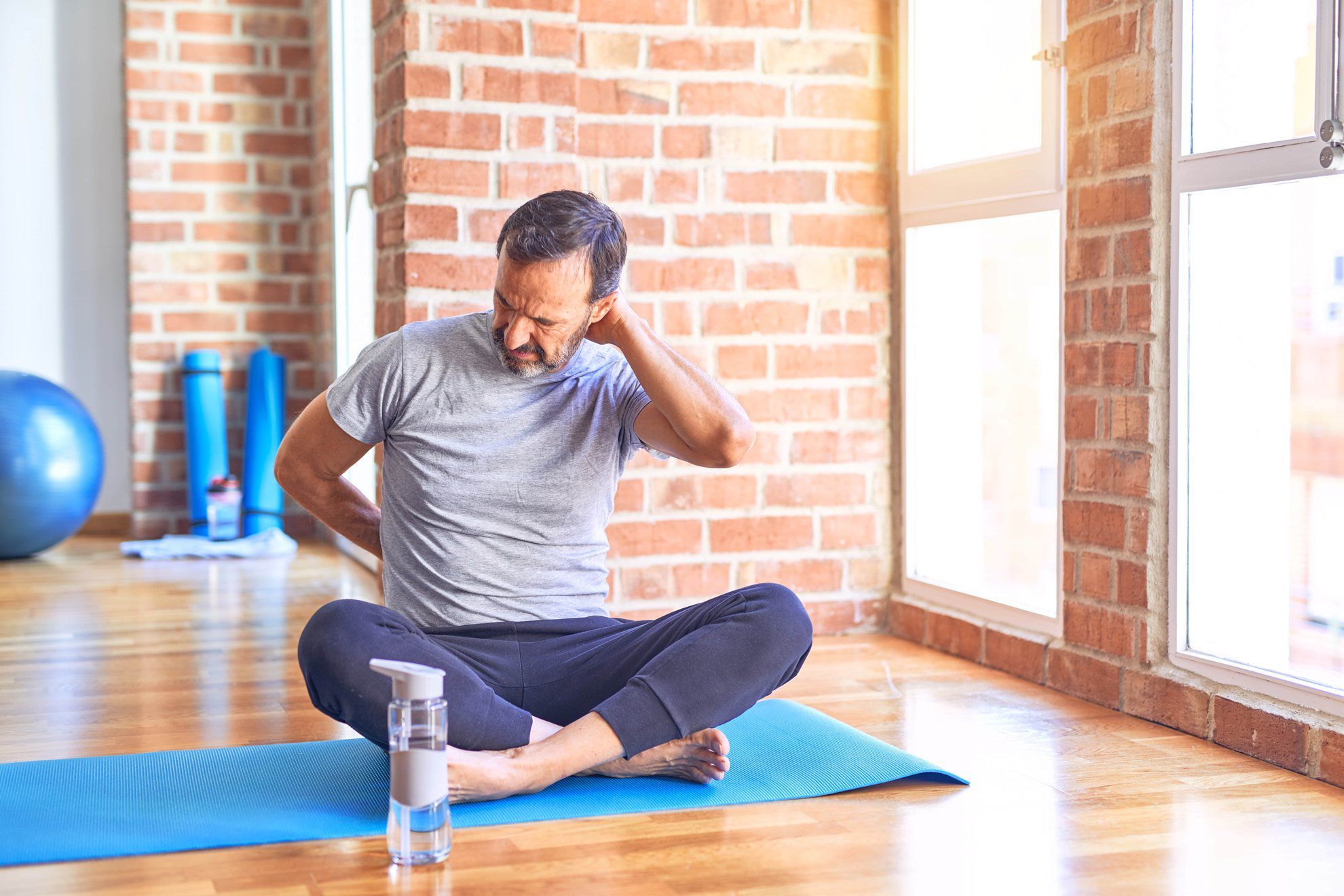 Sportivo di mezza età seduto sul tappetino mentre fa esercizi di stretching yoga in palestra. Soffre per un dolore al collo, si tocca il collo con la mano a causa di un dolore muscolare.